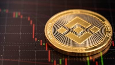 A close-up shot of a golden Binance Coin (BNB) resting on a dark digital screen displaying a cryptocurrency candlestick chart. The coin features the Binance logo and "BINANCE COIN" text. The chart shows red and green candlestick bars, indicating market fluctuations, against a black background with white grid lines. There's a subtle reflection of the chart on the coin's surface.