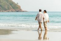 Happy Asian family senior couple walking together on tropical island beach at summer sunset. Retired elderly people relaxing and enjoy outdoor lifestyle travel nature ocean on holiday vacation.