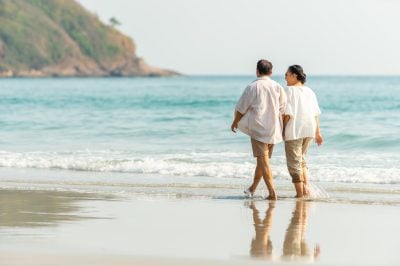 Happy Asian family senior couple walking together on tropical island beach at summer sunset. Retired elderly people relaxing and enjoy outdoor lifestyle travel nature ocean on holiday vacation.
