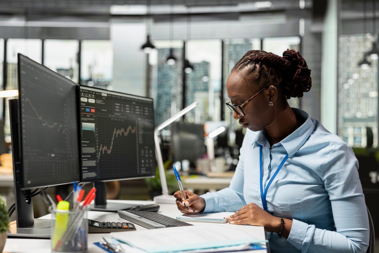 Risk analyst in office documents strategies while observing stock candle patterns on screen. Brokerage company worker reviews stock market shares volatility, recording insights in notepad