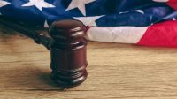 A gavel on a wooden table with a blurred American flag background, symbolizing the U.S. legal system.