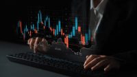 A close-up view of a person's hands typing on a black computer keyboard. Superimposed on the scene is a translucent digital financial chart, displaying blue and red candlestick bars, a white line graph, and various numerical figures, all set against a dark, blurred background.