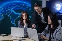 Four professionals gathered around laptops discussing an AI-related project, with digital brain graphics displayed on large screens in the background.