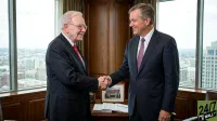 Warren Buffett, an older man with glasses and white hair, wearing a dark pinstripe suit and a red patterned tie, smiles warmly as he shakes hands with Greg Abel, a younger man with dark hair, wearing a dark suit and a purple patterned tie, who is also smiling. They stand in a modern office with large windows offering a view of a city skyline. On a polished wooden desk between them, there are open documents and a framed picture of a grand building. The mood is professional and amicable.