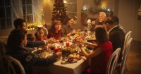 A large multi-generational family is gathered around a long dining table, celebrating a holiday meal in a warmly lit home decorated for the festive season. A roasted turkey is at the center of the table, surrounded by various dishes, wine glasses, and lit candles. Family members of different ages are smiling and conversing, enjoying the gathering, with a Christmas tree visible in the background.