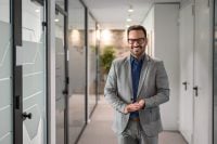 A smiling man in a grey suit, blue collared shirt, and black-rimmed glasses stands with his hands clasped, looking directly at the viewer. He has dark hair and a beard. He is in a modern office hallway with glass partitions on the left, featuring frosted geometric patterns, and white doors on the right. The hallway is brightly lit, with ceiling lights visible in the background.
