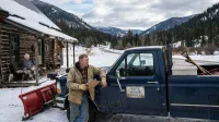 A wide shot of a snowy rural scene features a man in a work jacket and gloves leaning against a dark blue pickup truck fitted with a red snowplow on the left. The truck has a white, weathered sign reading 'RAY'S SENIOR SERVICES' on the door. Behind him, an older woman with white hair is bundled in a plaid blanket, sitting on a wooden bench on the porch of a rustic log cabin. In the background, a winding snowy road leads into pine-covered mountains under a cloudy sky.
