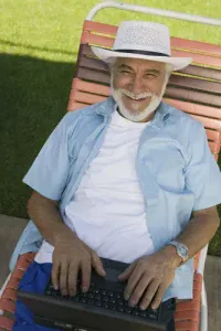 A smiling older man with a white beard and a white straw hat sits on an orange and brown striped lounge chair outdoors. He is wearing a light blue open shirt over a white t-shirt and blue shorts, with his hands resting on a black laptop in his lap. Green grass is visible in the background, bathed in sunlight.