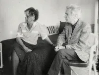 A black and white photo shows an older man and woman seated indoors. The woman, with light hair, sits on a dark patterned couch or bed, wearing a light collared shirt and dark skirt. She looks to the left, resting her left hand on the couch. To her right, an older man with white hair sits in a woven chair, wearing a plaid blazer and dark pants, also looking to the left. His hands are clasped in his lap.