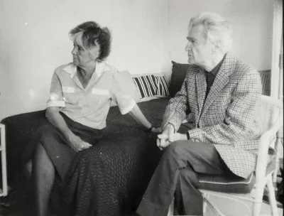 A black and white photo shows an older man and woman seated indoors. The woman, with light hair, sits on a dark patterned couch or bed, wearing a light collared shirt and dark skirt. She looks to the left, resting her left hand on the couch. To her right, an older man with white hair sits in a woven chair, wearing a plaid blazer and dark pants, also looking to the left. His hands are clasped in his lap.
