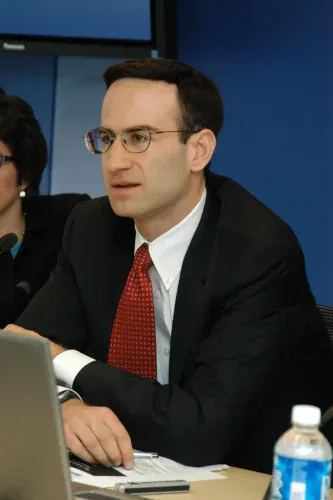 A man in a dark suit, white shirt, and a red patterned tie, wearing glasses, sits at a table. He is looking off to the left with an attentive expression. A silver laptop is in the foreground on the left, and a partial view of a plastic water bottle is on the right. Papers and a pen are visible on the table. A blue wall forms the background, and another person is partially visible on the far left.