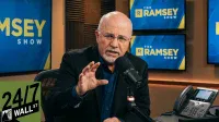 A bald man with a beard and glasses, wearing a dark suit jacket and a blue shirt, speaks intently into a black studio microphone. He is gesturing with his right hand. In the background, multiple blue screens display 'THE RAMSEY SHOW' in yellow text. A black desk phone is visible on the right, and a '24/7 WALL ST' logo is overlaid in the bottom left corner.