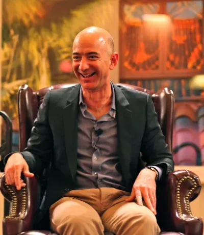 A bald man, likely Jeff Bezos, with a wide smile looks to the left while seated in a dark brown leather armchair. He is wearing a dark blazer over a light grey collared shirt with a small microphone clipped on, and light brown trousers. The background is softly blurred with warm, abstract patterns.