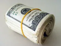 A close-up shot of a thick roll of U.S. one hundred dollar bills, secured tightly by a bright yellow rubber band. The bills are slightly fanned on one end, revealing their edges, and the green and white details of the currency are clearly visible, including parts of the serial number and Federal Reserve seal.