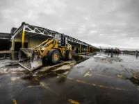 A large yellow Caterpillar front-end loader with a wide bucket is parked on a wet, dark paved surface with faint yellow markings. In the background, a long, partially deconstructed industrial building with white walls and yellow support beams stretches under a cloudy, grey sky. The ground shows reflections from puddles.