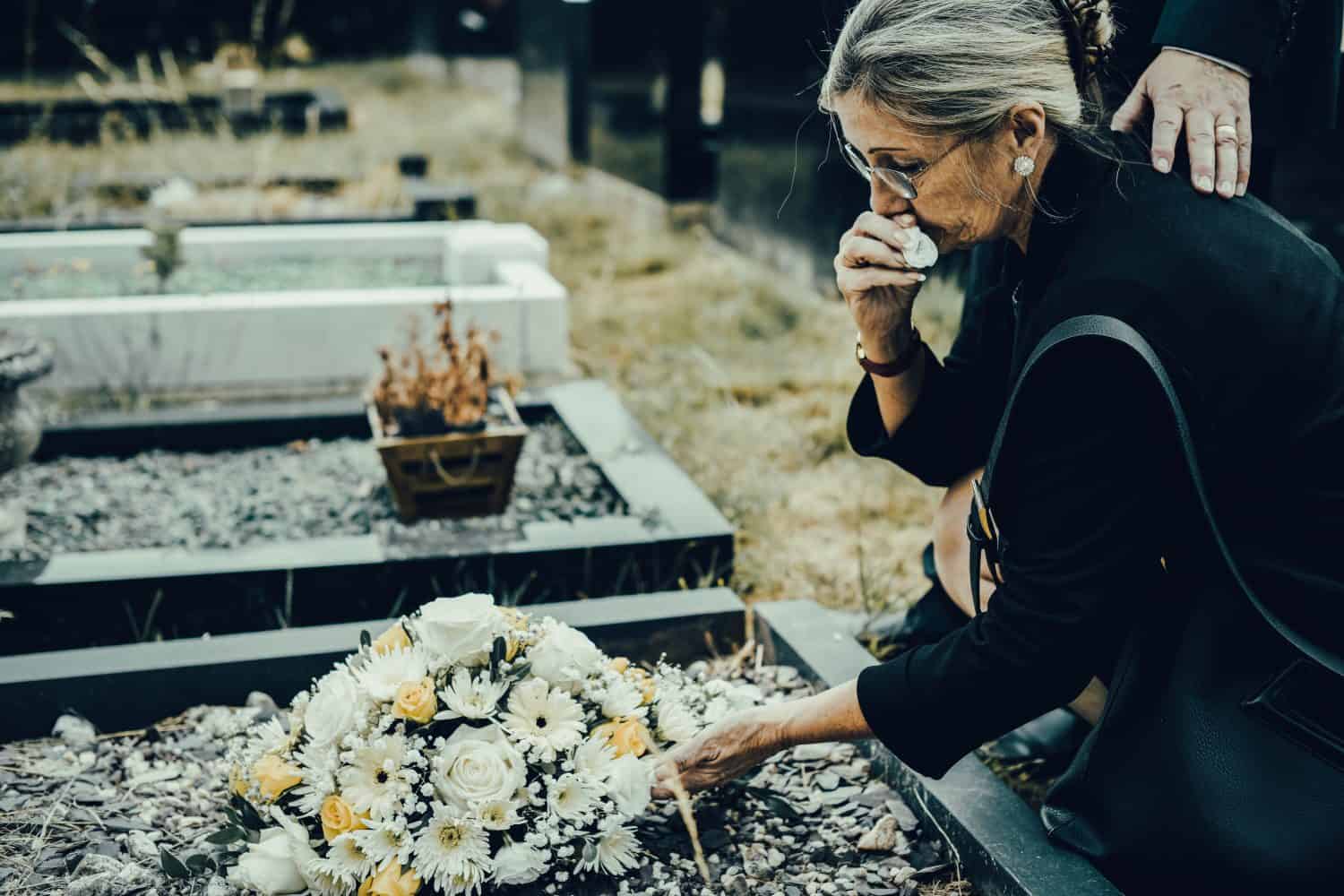 Elderly woman mourning at a grave, placing flowers. Mourning woman in black, showing grief in funeral. Elderly woman at cemetery, expressing sorrow and loss. Funeral event photography concept.