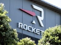A close-up shot of the Rocket Lab company logo and name on a dark grey building facade. The logo features a large white stylized 'R' symbol with a red arrow-like shape pointing left next to it, and a small white triangle beneath the 'R'. Below the logo, the word 'ROCKET' is spelled out in bold, white, 3D letters. Green bushes with sunlit leaves partially cover the lower part of the sign and building.