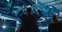 View from behind of a man in a dark suit with arms raised in excitement or reaction, standing before a large, curved wall of glowing computer screens displaying various financial charts and data in blue, green, and red hues. A woman is partially visible on the right, also looking at screens. The overall mood is intense and dynamic in the dark, high-tech control room.