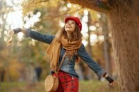 Hello autumn. smiling elegant 40 years old woman in jeans shirt and red hat with autumn leaf, scarf and bag rejoicing in the city park.