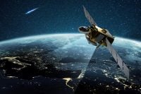 A gold and gray communication satellite with large rectangular solar panels orbits high above Earth at night. A bright beam of light emanates from the satellite, illuminating a segment of the continents below, where city lights glow. In the dark, star-filled sky, a luminous blue-streaked meteor or comet is visible in the upper left.