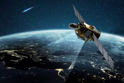 A gold and gray communication satellite with large rectangular solar panels orbits high above Earth at night. A bright beam of light emanates from the satellite, illuminating a segment of the continents below, where city lights glow. In the dark, star-filled sky, a luminous blue-streaked meteor or comet is visible in the upper left.