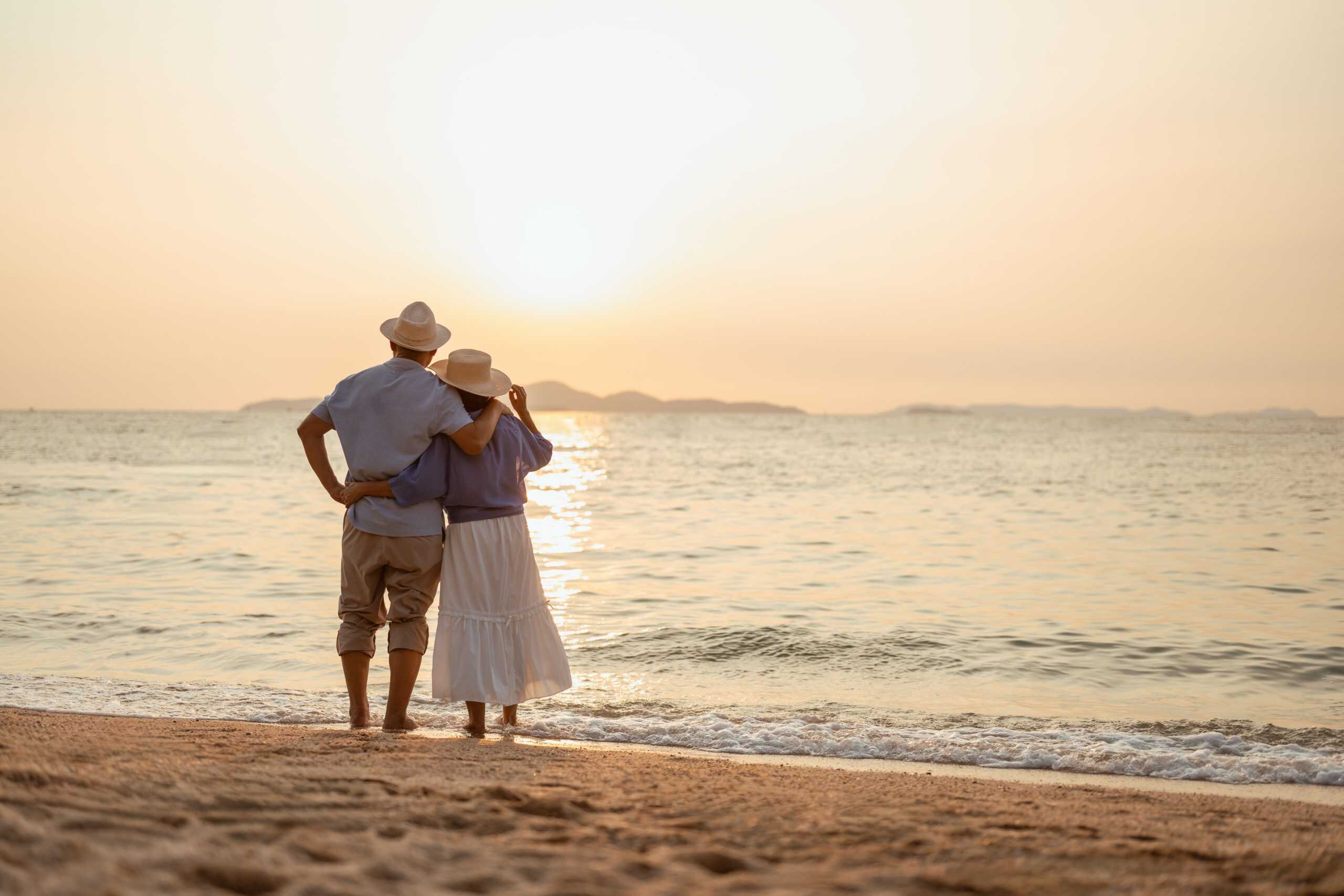 retired, retirement, couple, senior, elderly, beach, recreation, insurance, planning, vacation. A couple standing on the beach at sunset, with the man holding the woman's hand.