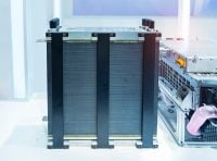 A tall, rectangular industrial fuel cell stack, featuring numerous thin horizontal layers and secured by four vertical black support straps, stands in a brightly lit, clean environment. A silver metallic top plate with several black cylindrical knobs is visible at the apex. To the right, a portion of another silver industrial unit with a bright orange electrical connector is visible.