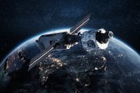 An overhead view of a large, complex satellite with extended solar panels, orbiting high above Earth at night. Bright clusters of golden city lights are visible across the dark landmasses below, with a prominent blue atmospheric glow along the curved horizon. Numerous white stars dot the dark blue background of space.