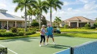 An older man and woman are smiling on a green pickleball court, each holding a blue pickleball paddle. The woman wears a purple shirt and black capris, and the man wears a blue shirt and grey shorts. Behind them are lush green lawns, several tall palm trees, and two single-story houses with light-colored exteriors and different roof styles under a bright blue sky with scattered clouds. A green golf cart is parked in the background near one of the houses.