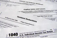 A close-up, angled view of multiple white U.S. federal income tax forms with black printed text, overlapping and fanned out. Clearly visible headings include 'Form 1040 U.S. Individual Income Tax Return,' 'Itemized Deductions,' 'Capital Gains and Losses,' and 'Interest and Ordinary Dividends.' The forms are pristine and show detailed instructions and legal language.