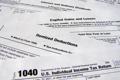 A close-up, angled view of multiple white U.S. federal income tax forms with black printed text, overlapping and fanned out. Clearly visible headings include 'Form 1040 U.S. Individual Income Tax Return,' 'Itemized Deductions,' 'Capital Gains and Losses,' and 'Interest and Ordinary Dividends.' The forms are pristine and show detailed instructions and legal language.