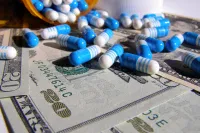 A close-up shot shows a pile of blue and white medical capsules spilled from an orange medicine bottle onto several US twenty-dollar bills. The capsules are striped or solid blue, and some bills are partially visible, showing '20' and serial numbers like 'JF03447614C'.
