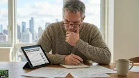 A middle-aged man with gray hair and glasses, wearing a brown knitted sweater, sits at a wooden table. He holds a pen in his right hand, writing in a spiral-bound notebook, while his left hand rests on his chin in a thoughtful pose. To his left, a black tablet displays an article titled 'Manhattan Luxury Rentals Surge: Top Leases Hit $100k+/Month' with a blue line graph. Papers are scattered on the table, and a light brown mug is visible on the right. In the background, a large window reveals a cityscape with tall buildings under a bright sky.