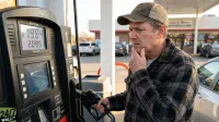 A man with a pensive expression, wearing a brown and black plaid shirt and a worn baseball cap, stands at a gas pump, holding the black fuel nozzle. His right hand is touching his chin. The gas pump's digital display shows '119.84' at the top and '2.095 PER GALLON' below. A convenience store and other vehicles are visible in the sunny background.