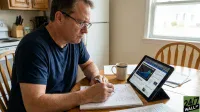 A middle-aged man with glasses and a dark blue t-shirt sits at a wooden table, intently writing in a spiral notebook with a pen. To his right, a white mug with blue stripes sits next to a tablet displaying a financial news website with stock charts and headlines about 'Big Tech AI' and 'depreciation'. A kitchen area with light wood cabinets and a white refrigerator is visible in the background, along with a window showing an outdoor scene.