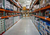 A long, brightly lit aisle inside a large warehouse store extends into the distance. Tall, orange and green industrial metal shelves on both sides are stacked high with numerous white and blue cardboard boxes containing various consumer goods, including microwaves, some resting on wooden and plastic pallets. The concrete floor is clean and reflective, and the ceiling has exposed beams and industrial lighting.
