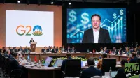 A large conference room features a U-shaped table where numerous dignitaries are seated. On the left screen, the G20 logo is displayed, and a man stands at a podium. On the right, a large screen shows a close-up of Elon Musk speaking, with dollar signs and an upward-trending bar graph in the background. The room is well-lit, and flags from various countries are visible on the tables.