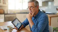 A man with grey hair and glasses sits at a kitchen table, holding a tablet that displays financial charts and graphs. He wears a blue button-up shirt and has his right hand resting on his chin, looking intently at the screen with a thoughtful or concerned expression. To his left, a white coffee mug and a folded newspaper are on the wooden table. The background shows a brightly lit modern kitchen with white cabinets and a dishwasher.
