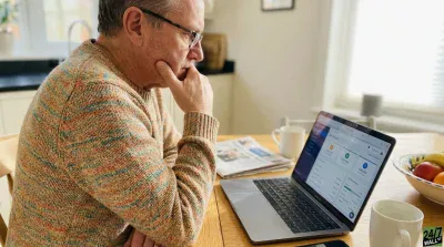 A thoughtful older man wearing glasses and a multi-colored knitted sweater sits at a wooden table, looking intently at a silver laptop screen. His right hand is raised to his chin in a pensive gesture. On the table, next to the laptop, are a folded newspaper, a white mug, and a bowl of fruit. The background shows a bright, softly lit kitchen or dining area with a window.
