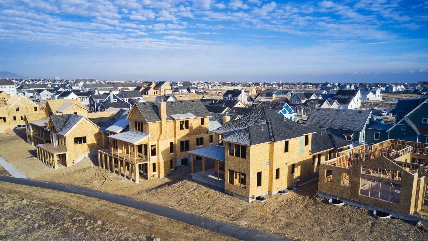 An aerial view shows a new housing development with many single-family homes under construction. In the foreground, homes are framed with light brown wood sheathing, some with dark gray roofs, and dirt patches surround them. Further back, numerous completed and partially completed homes with colorful siding (blue, gray, white) extend into the distance under a bright blue sky with scattered white clouds, with faint mountains on the horizon.