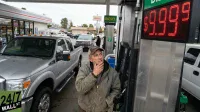 A man in a baseball cap and work jacket stands at a silver diesel gas pump, looking up with a thoughtful expression at the red LED price display. The display prominently shows '$9.999'. A silver Ford pickup truck is parked to the left of the pump. In the background, a convenience store and a 'DIESEL' sign are visible under an overcast sky.