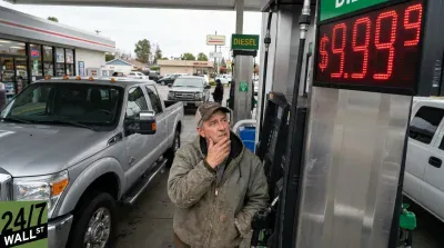 A man in a baseball cap and work jacket stands at a silver diesel gas pump, looking up with a thoughtful expression at the red LED price display. The display prominently shows '$9.999'. A silver Ford pickup truck is parked to the left of the pump. In the background, a convenience store and a 'DIESEL' sign are visible under an overcast sky.