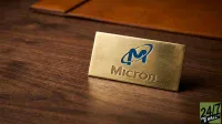 A close-up shot of a rectangular, brushed gold nameplate bearing the blue and white Micron Technology logo and the word 'Micron' embossed in gold. The nameplate is positioned on a dark brown wooden desk. A blurred brown leather desk accessory is visible in the background.