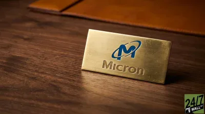 A close-up shot of a rectangular, brushed gold nameplate bearing the blue and white Micron Technology logo and the word 'Micron' embossed in gold. The nameplate is positioned on a dark brown wooden desk. A blurred brown leather desk accessory is visible in the background.