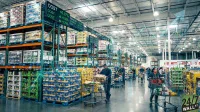 A wide-angle photograph inside a large, brightly lit warehouse store, reminiscent of Costco. Numerous shoppers push silver shopping carts down wide aisles flanked by towering green and blue metal shelving units packed with a variety of bulk packaged goods. On the left, a digital black screen displays "COST" three times, each followed by green text indicating percentage increases like "+0.77%", "+0.65%", and "+1.79%", accompanied by green upward arrows. A faint, translucent blue arrow points diagonally upwards from the lower left to the upper right of the image, suggesting growth.