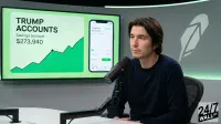 A man with dark hair and a dark blue long-sleeved shirt sits at a white table in front of a microphone, looking to his right. Behind him, a large screen displays 'TRUMP ACCOUNTS' and '$273,940' with an upward-trending green graph, alongside a smartphone screen showing an 'Investing' app with similar financial data. A feather-like logo is visible on the right side of the screen, and a '24/7 WALL ST' logo is in the bottom right corner of the image.