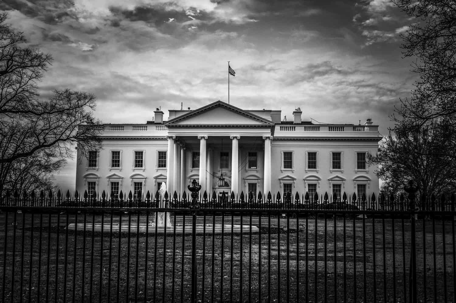 Black and white image of the White House presidential residence and office in Washington DC with iron fence and fountain in front and cloudy sky in background