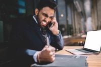 Furious nervous businessman biting bullet dressed in black suit white shirt tie surfing netbook making call on cellphone punching on wooden table in city cafe in summer