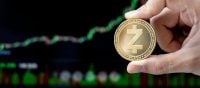 A human hand holds a shiny gold Zcash cryptocurrency coin, featuring the stylized 'Z' logo and text like 'ZCASH', 'decentralized', 'shielded', and 'secured'. The background is a blurry dark screen displaying a financial market chart with fluctuating green and red lines and bars.