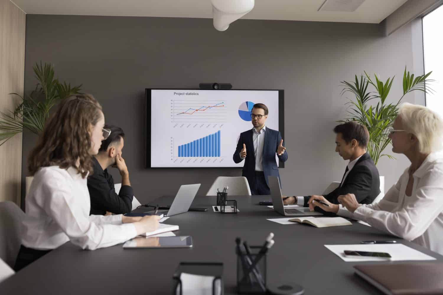 Male coach hold seminar for staff, makes presentation, provides sales statistics explanation gathered in modern conference room, with monitor screen with infographic charts, graphs and diagrams data